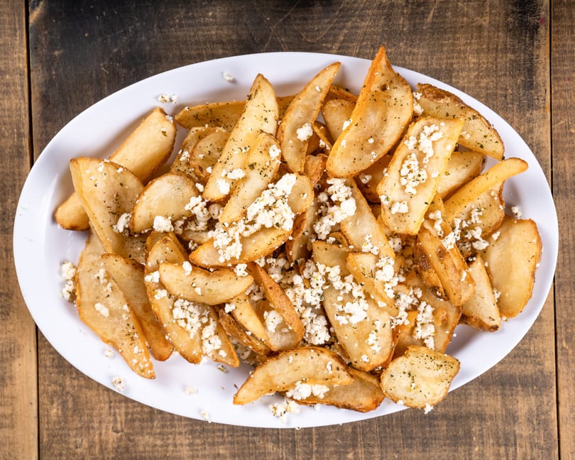 Golden potato wedges topped with crumbled feta cheese and herbs on a white platter.