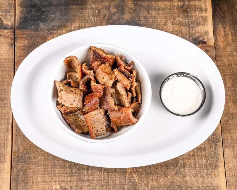 A white bowl filled with gyro meat and a small cup of tzatziki sauce on a white plate.
