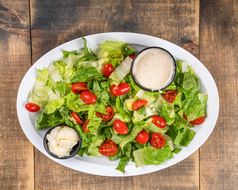 Fresh green salad with cherry tomatoes, dressing, and shaved cheese on a white platter.