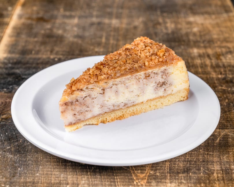 Slice of cheesecake with a crumbly nut topping on a white plate, on a rustic wooden table.