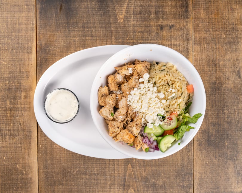 Chicken and rice bowl with feta, salad, and a side of dressing on a wooden table.