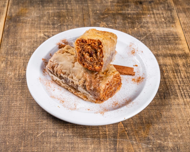 Two pieces of baklava, one cut open, on a white plate with cinnamon sticks and powder.