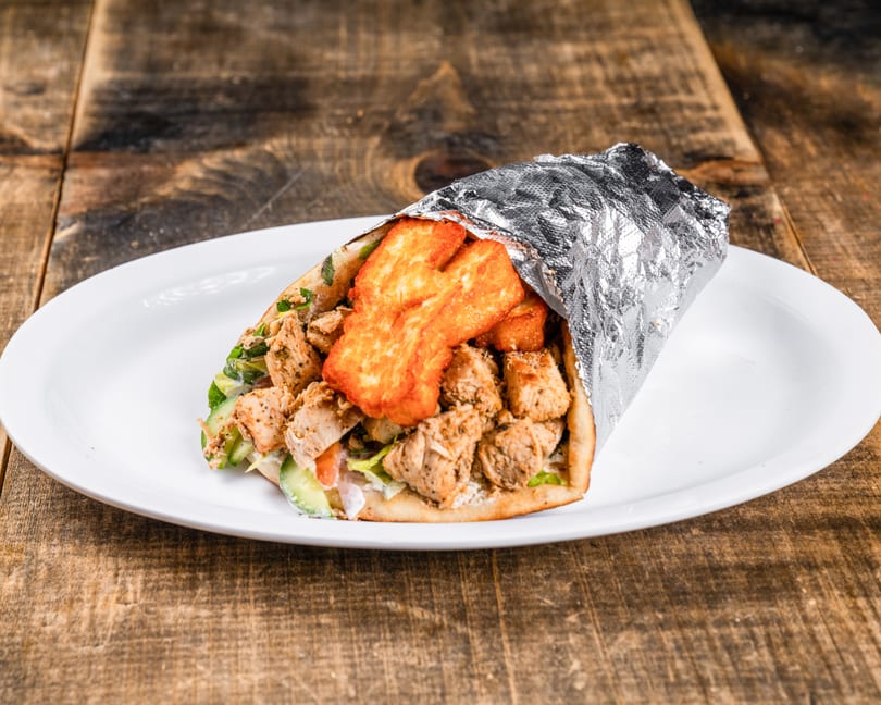Chicken and halloumi wrap, partially wrapped in foil, on a white plate on a wooden table.