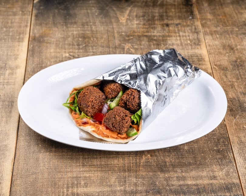 Falafel wrap with lettuce, tomatoes, onions, and sauce, partially wrapped in foil on a white plate.
